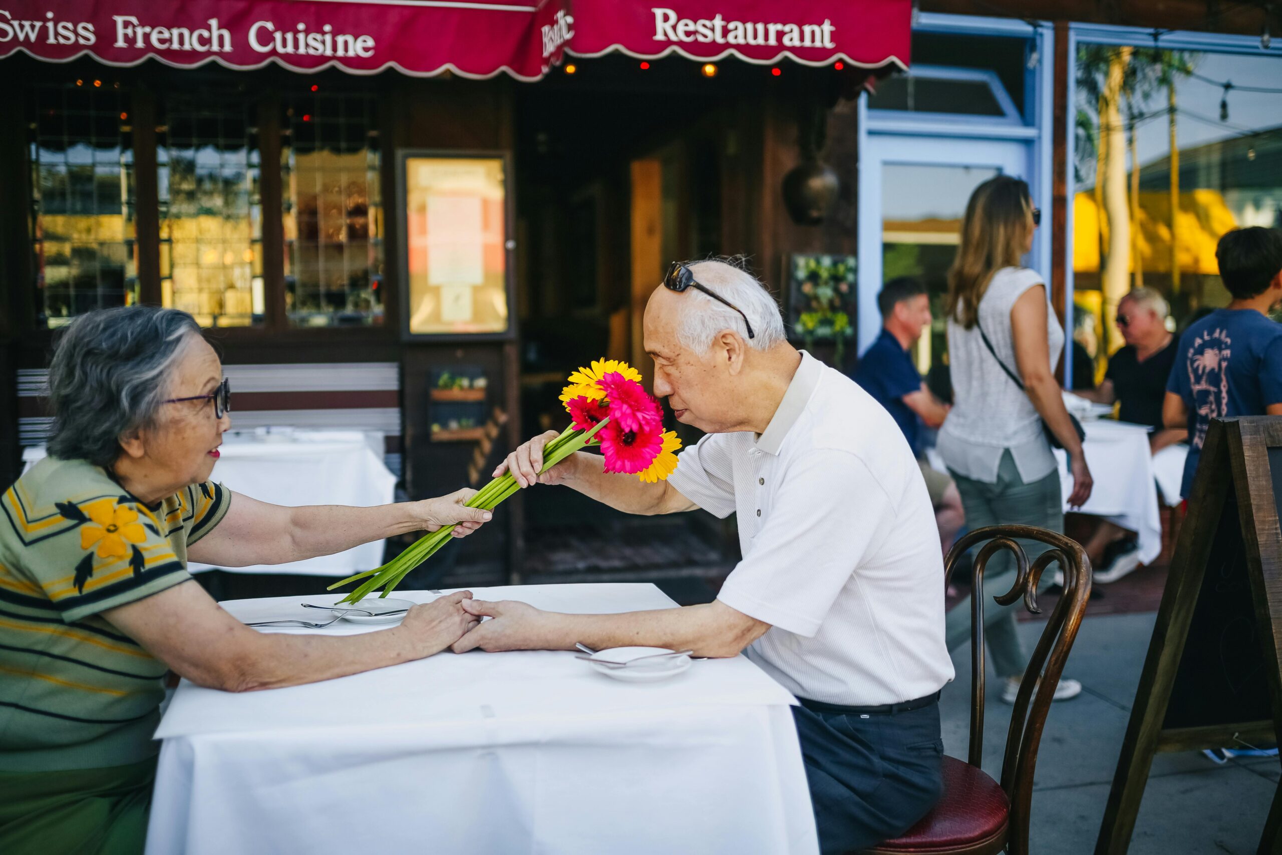 An elderly couple enjoys a heartfelt moment at an outdoor cafe, exchanging colorful flowers.